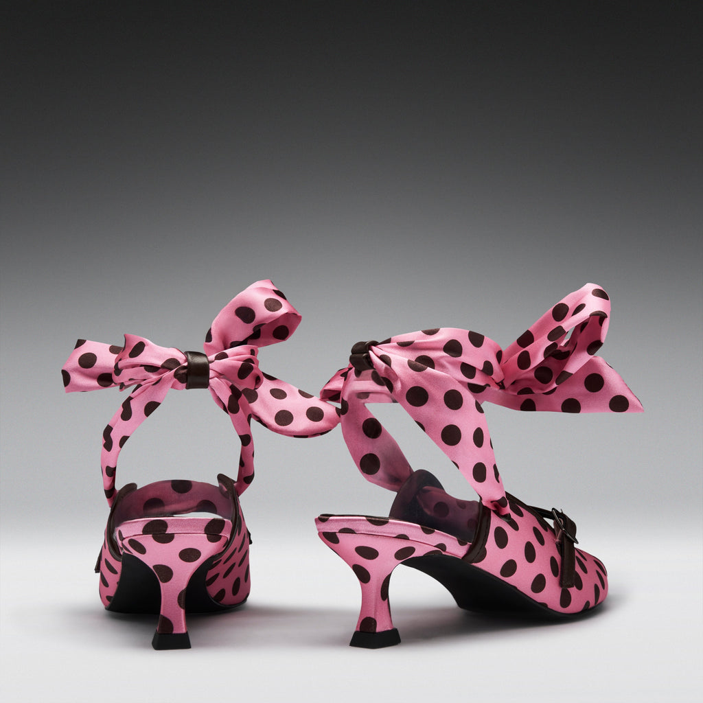 A pair of pink satin high-heeled sandals with black polka dots and large matching bow straps, shown from the back on a dark-to-light gray gradient studio background.