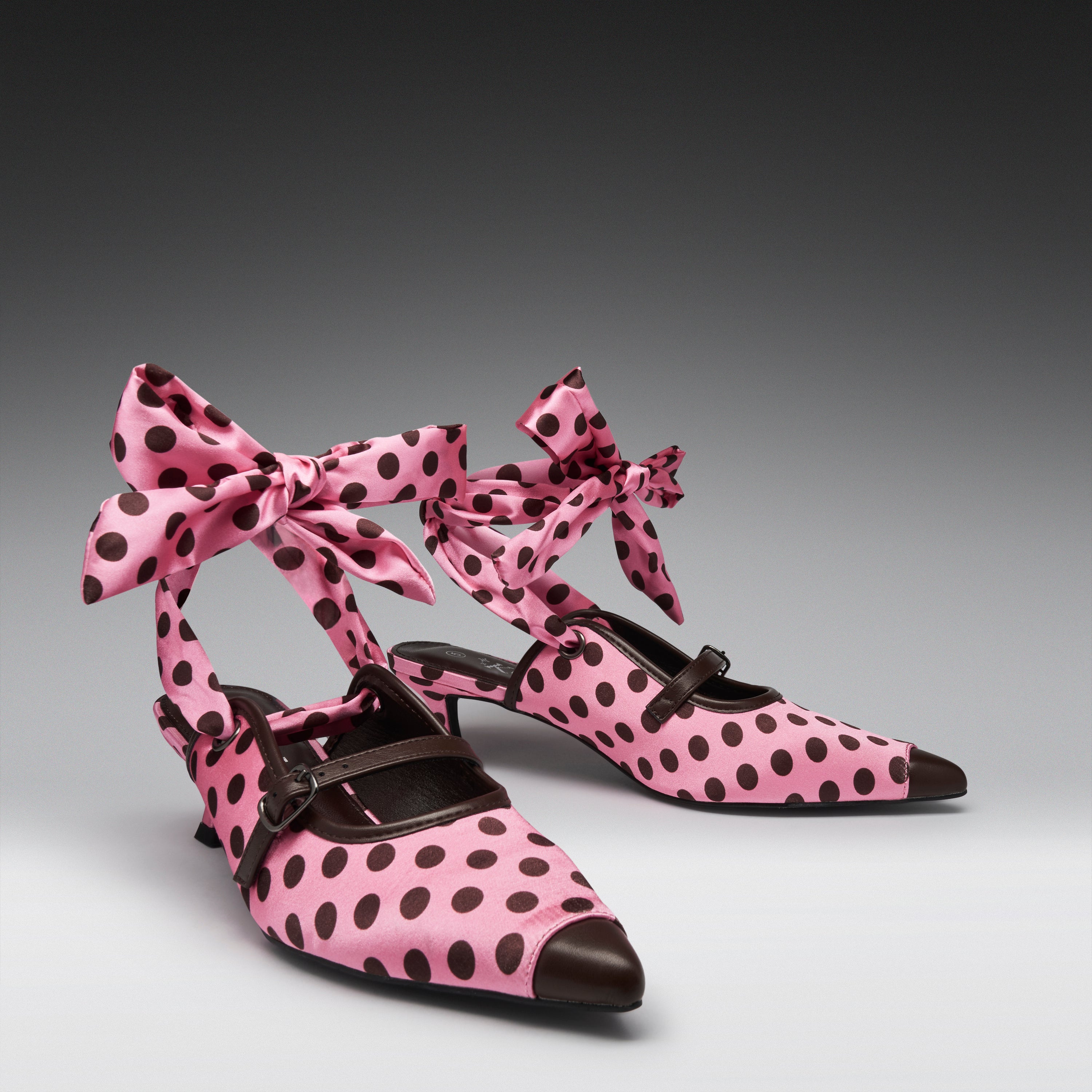 Pink pointed-toe Mary Jane-style heels with dark brown polka dots, bow ankle straps, and dark brown leather trim, photographed on a gray gradient studio background.