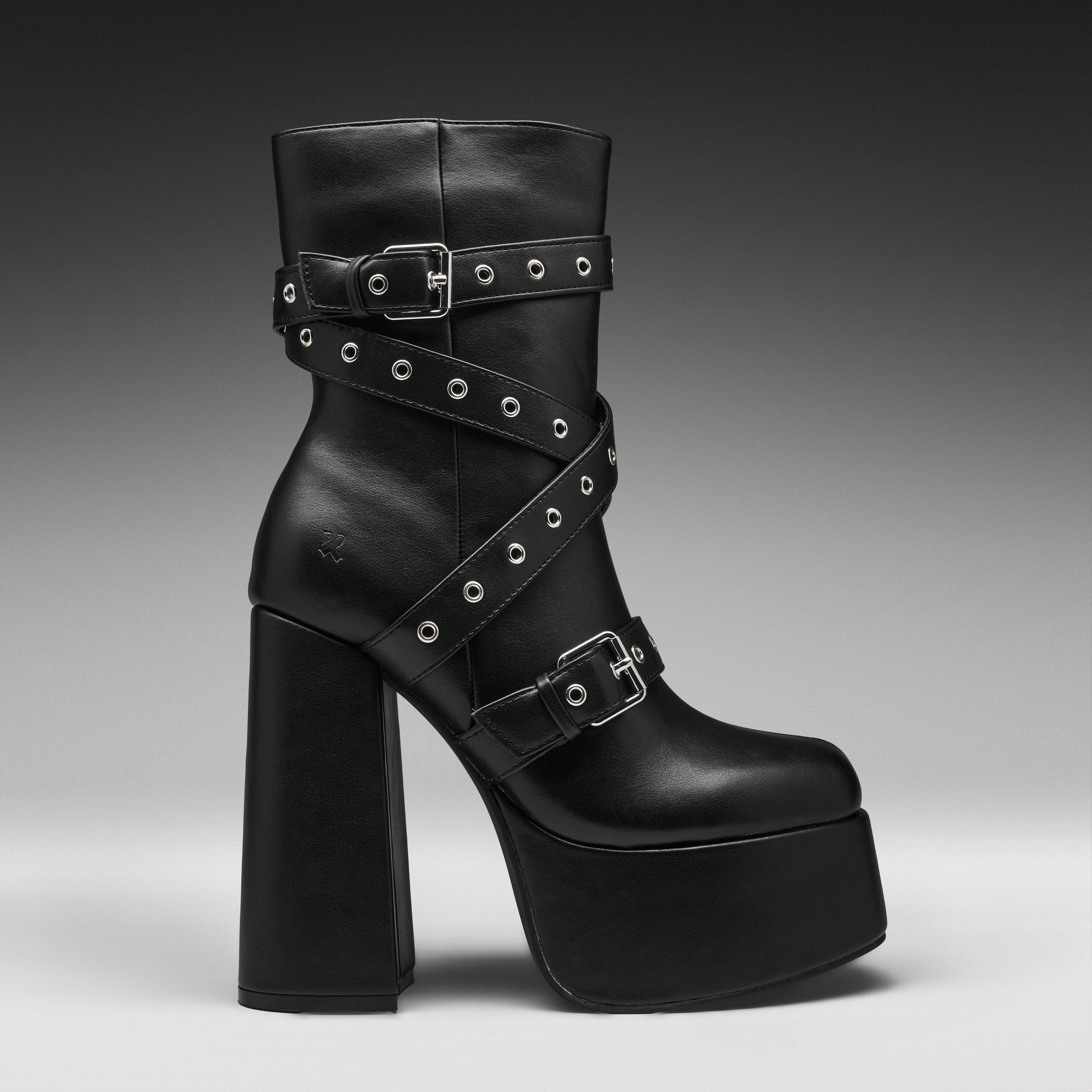 A black leather ankle boot with a chunky block heel, platform sole, and multiple decorative straps adorned with silver studs and buckles, shown against a dark gray gradient background.