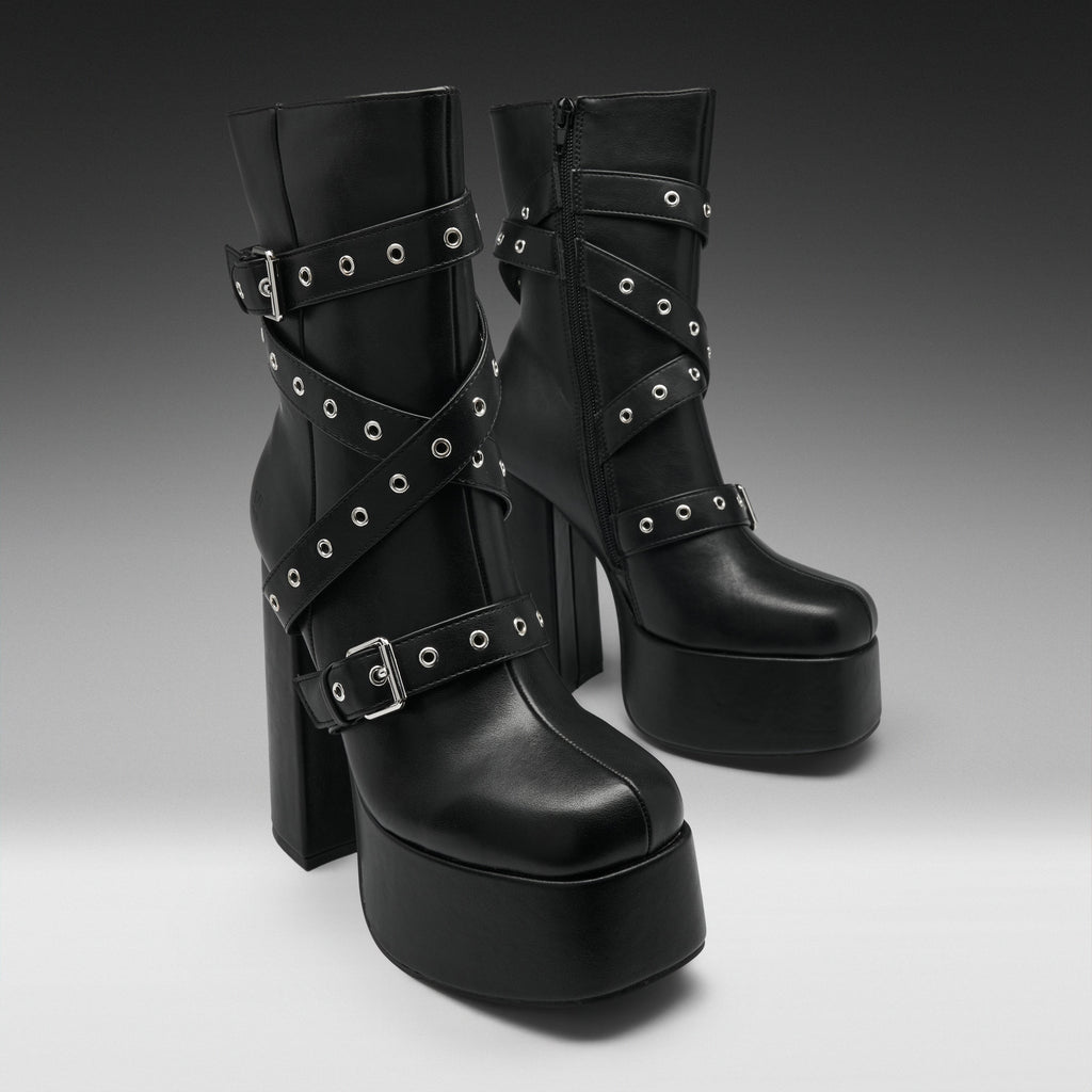 A pair of black platform ankle boots with high block heels, decorated with multiple crisscrossing straps and silver-tone metal grommets, shown against a dark-to-light gray gradient studio background.