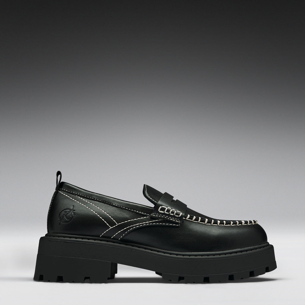A single black leather loafer with a chunky lug sole, featuring contrast white stitching and a decorative strap across the vamp, shown against a dark gray gradient background.