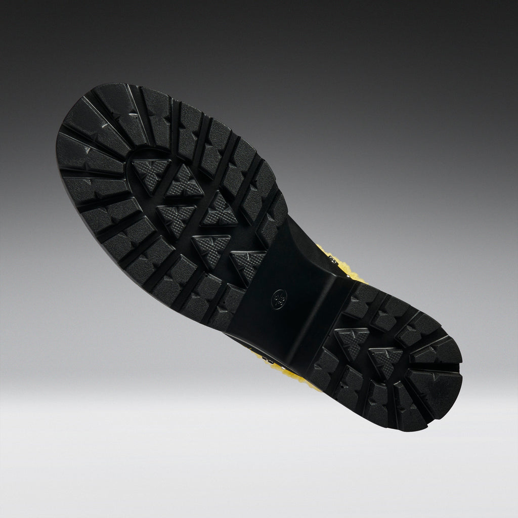 A close-up of a black rubber boot sole with deep tread patterns, shown against a dark gray to light gray gradient background.