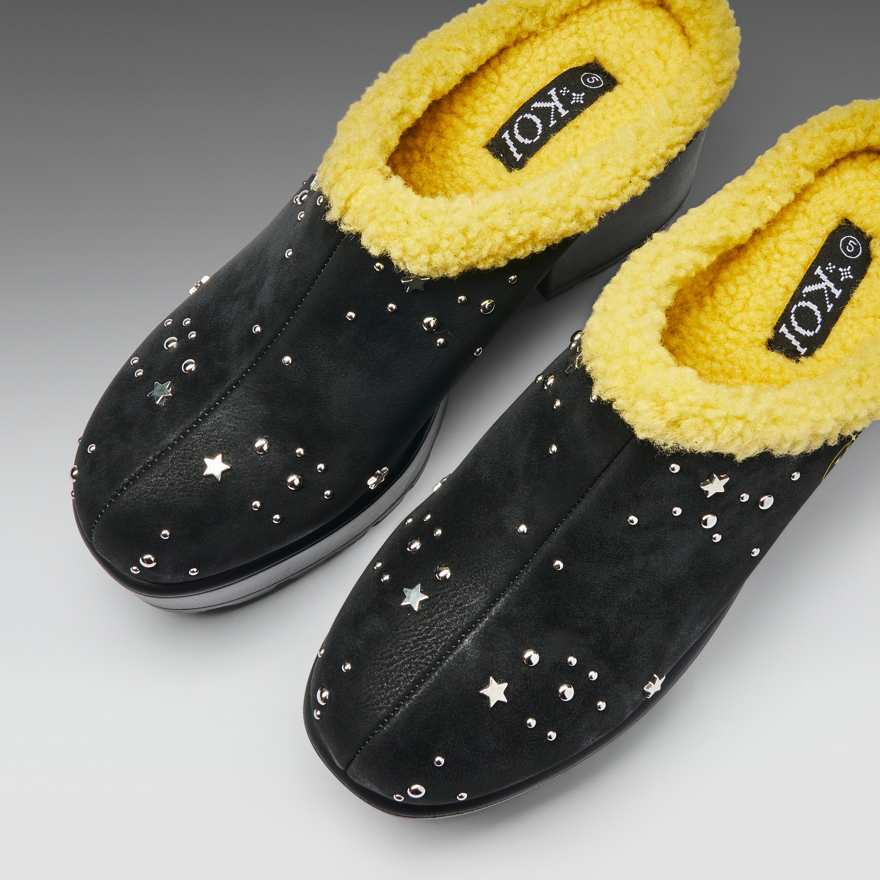 A pair of black slip-on mule-style slippers with bright yellow sherpa lining and scattered silver star-shaped studs, shown on a neutral gray background.