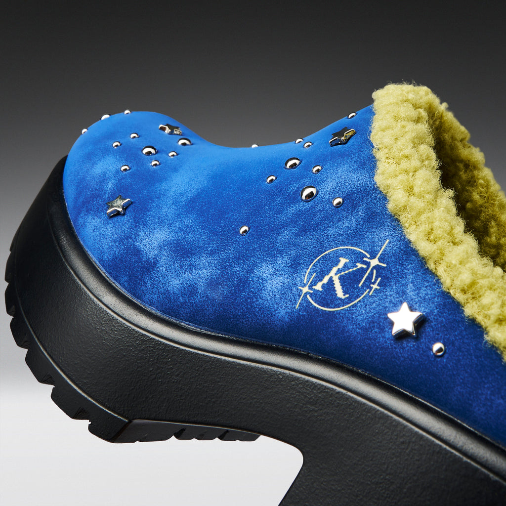 A close-up of a blue suede clog-style shoe with a black platform sole, a yellow-green sherpa lining, and silver star and stud embellishments.