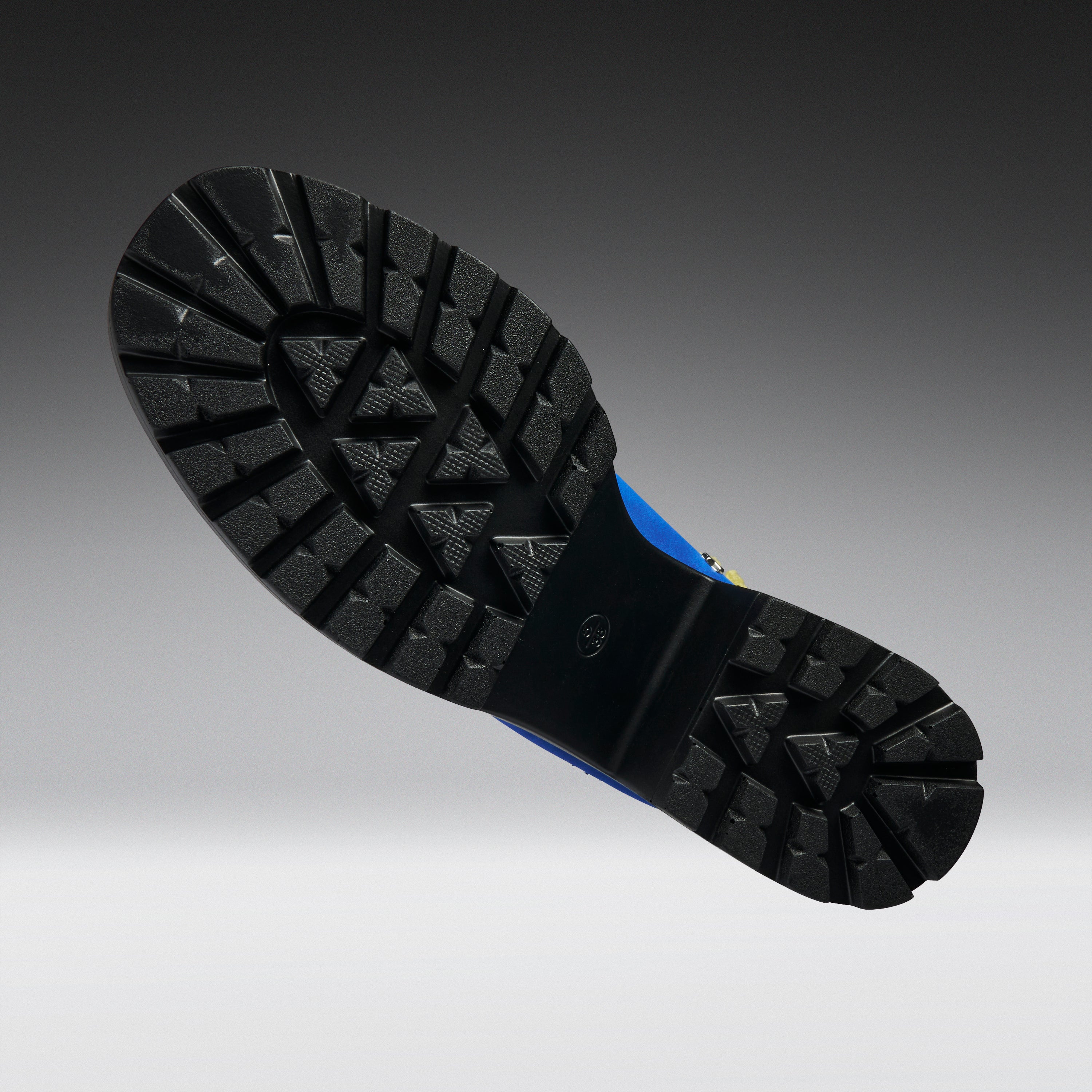 A close-up of a black rubber boot sole with deep tread patterns, shown against a dark gray to light gray gradient background.
