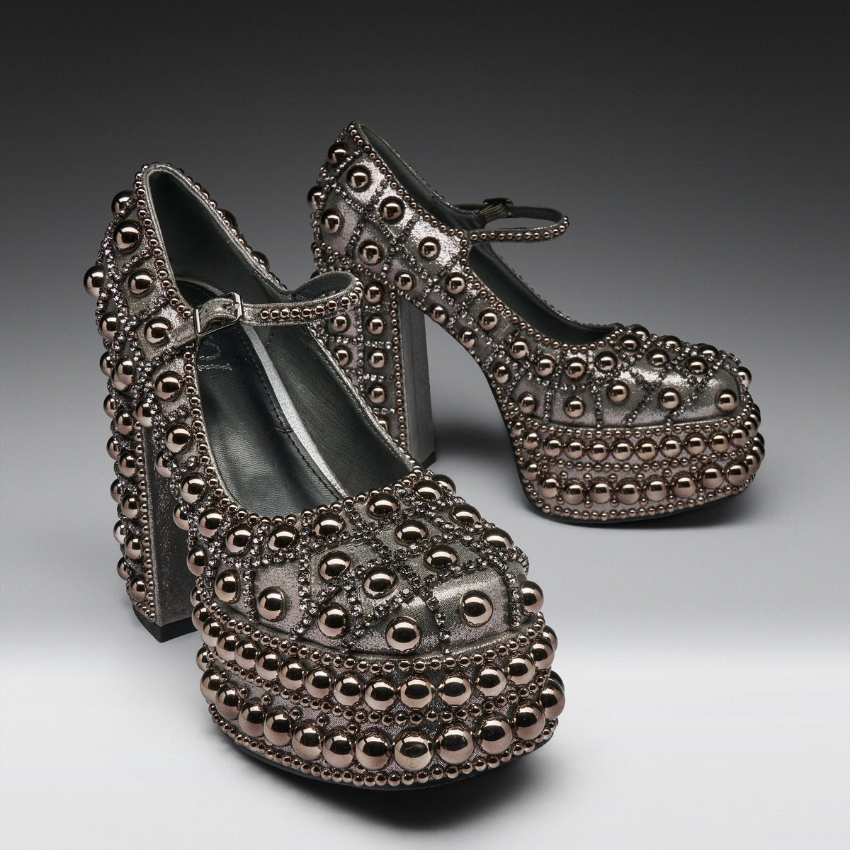 A pair of metallic silver platform Mary Jane shoes heavily embellished with rows of dark metallic studs and beads, set against a dark gray studio background.