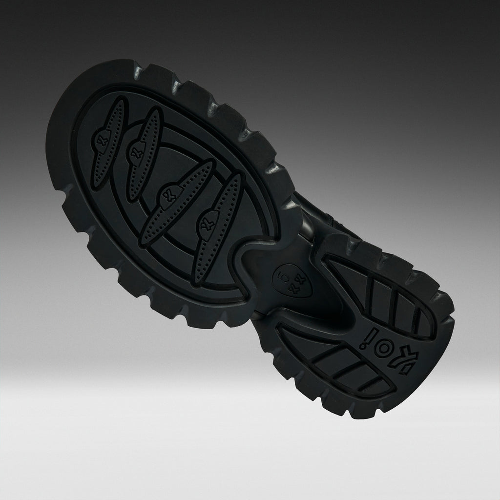 A close-up studio shot of a black rubber shoe sole, showing the tread pattern and embossed branding, set against a dark gray gradient background.