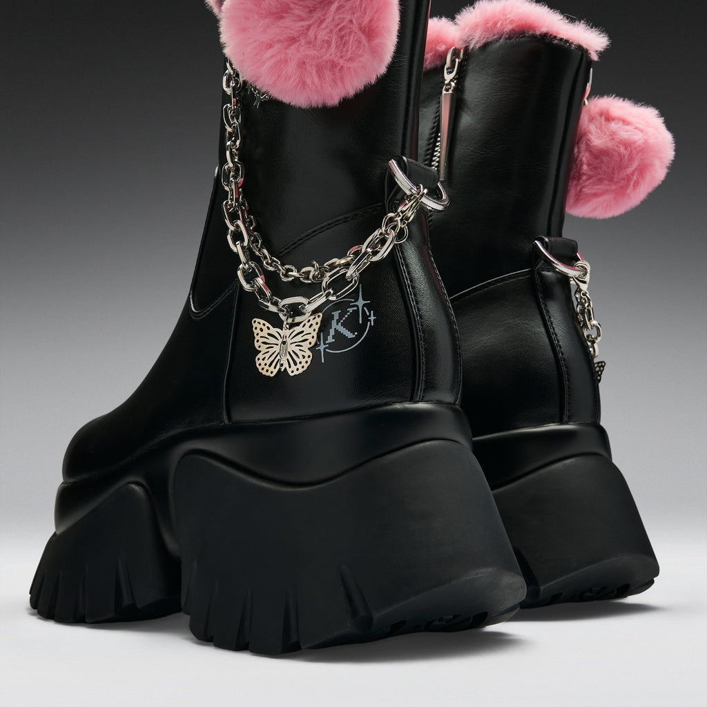 Black leather ankle boots with chunky lug soles, pink faux-fur trim, silver chain details, and a gold butterfly charm, shown on a dark gray studio background.