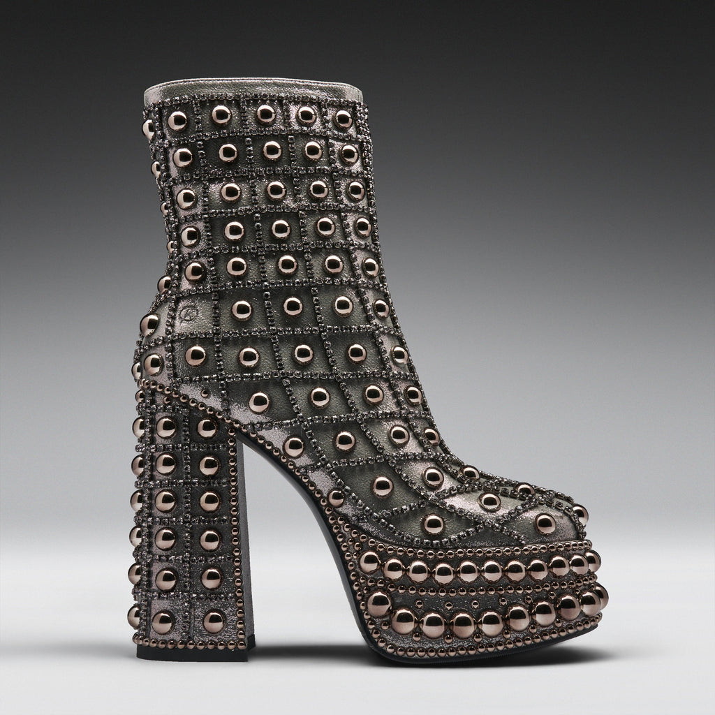A single high-heeled platform ankle boot covered in rows of metallic studs and rhinestones, shown in profile against a dark gray gradient studio background.