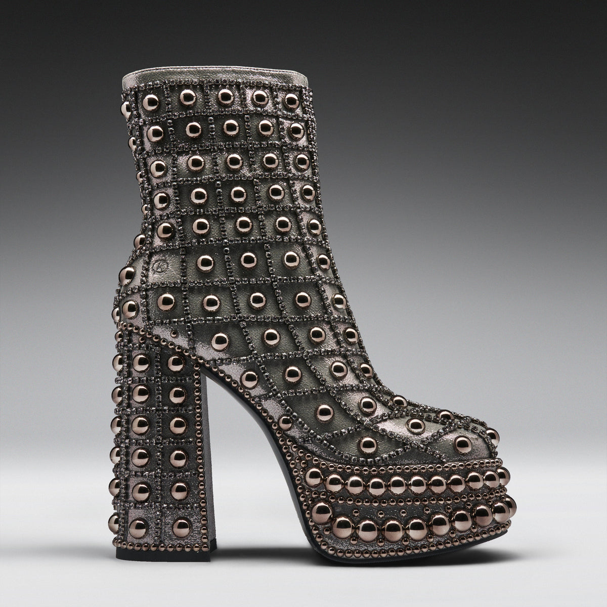 A single high-heeled platform ankle boot covered in rows of metallic studs and rhinestones, shown in profile against a dark gray gradient studio background.