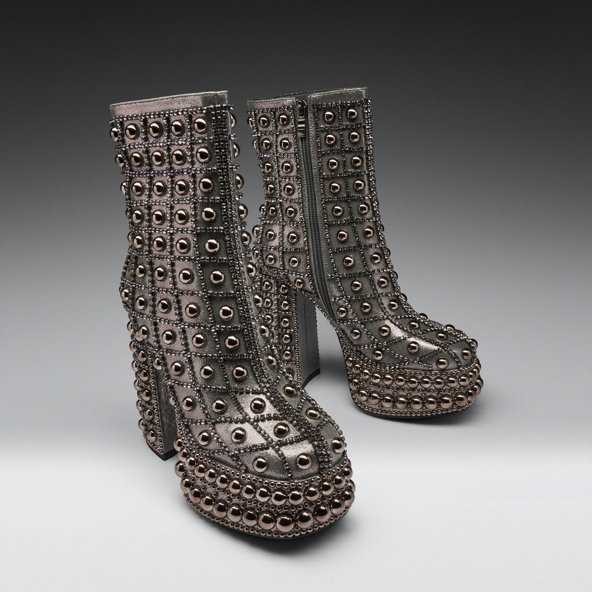 A pair of metallic silver platform ankle boots heavily embellished with rows of round dark metallic studs and rhinestones, set against a dark-to-light gray studio background.