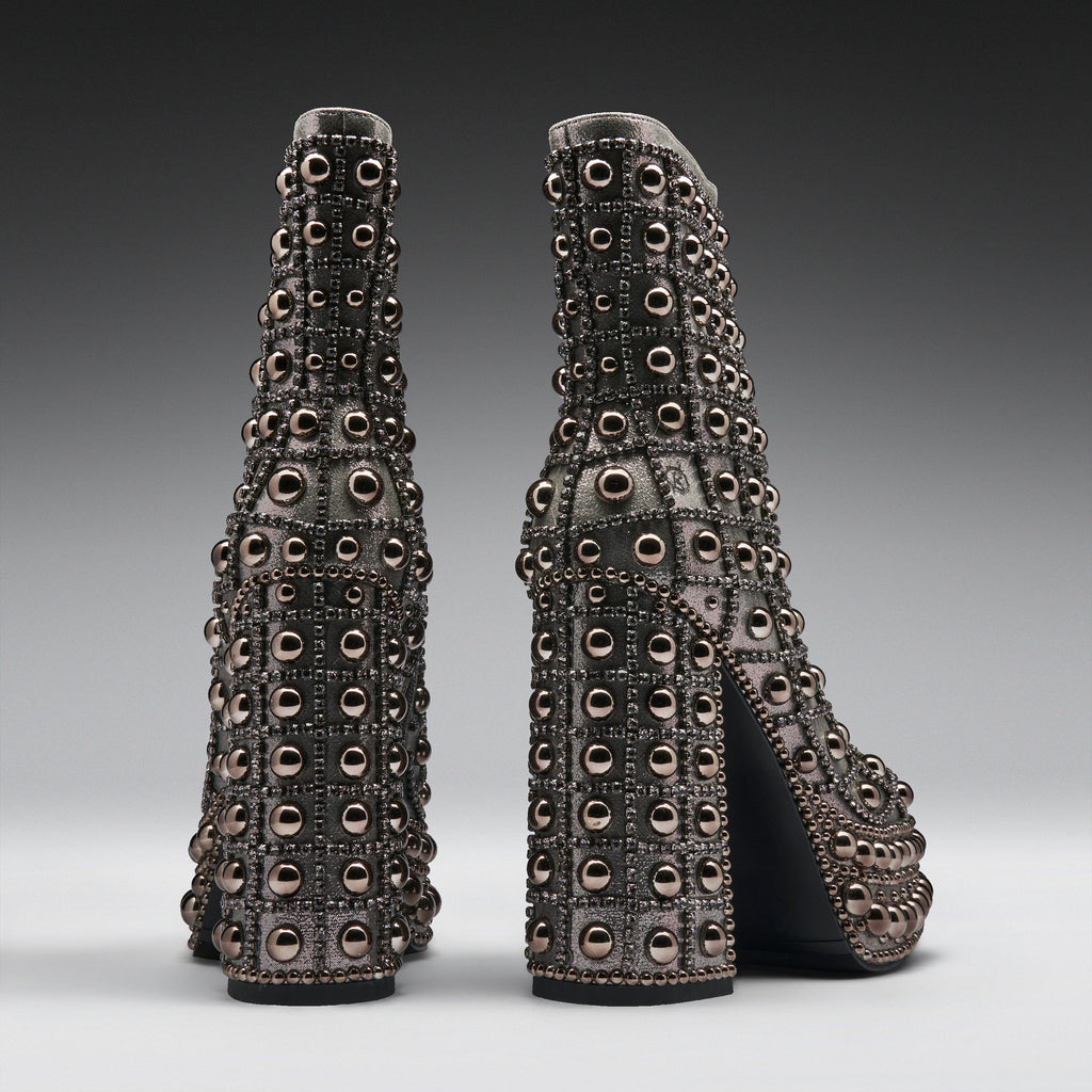 A pair of high-heeled boots with chunky block heels, covered in rows of metallic silver studs and circular embellishments, shown from the back on a dark gray gradient background.