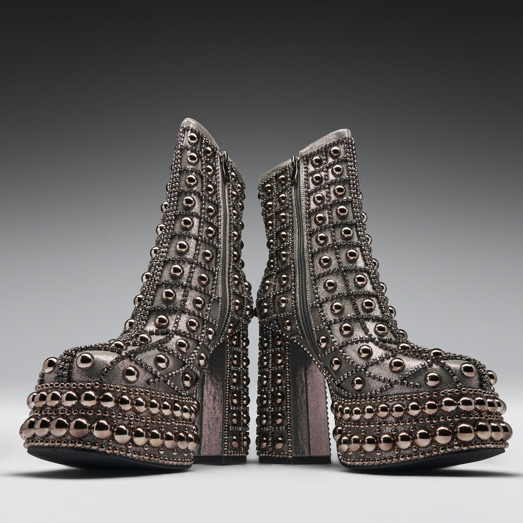 A pair of heavily embellished platform ankle boots with metallic rose gold studs and rhinestone detailing, shown from the side against a dark gray gradient background.