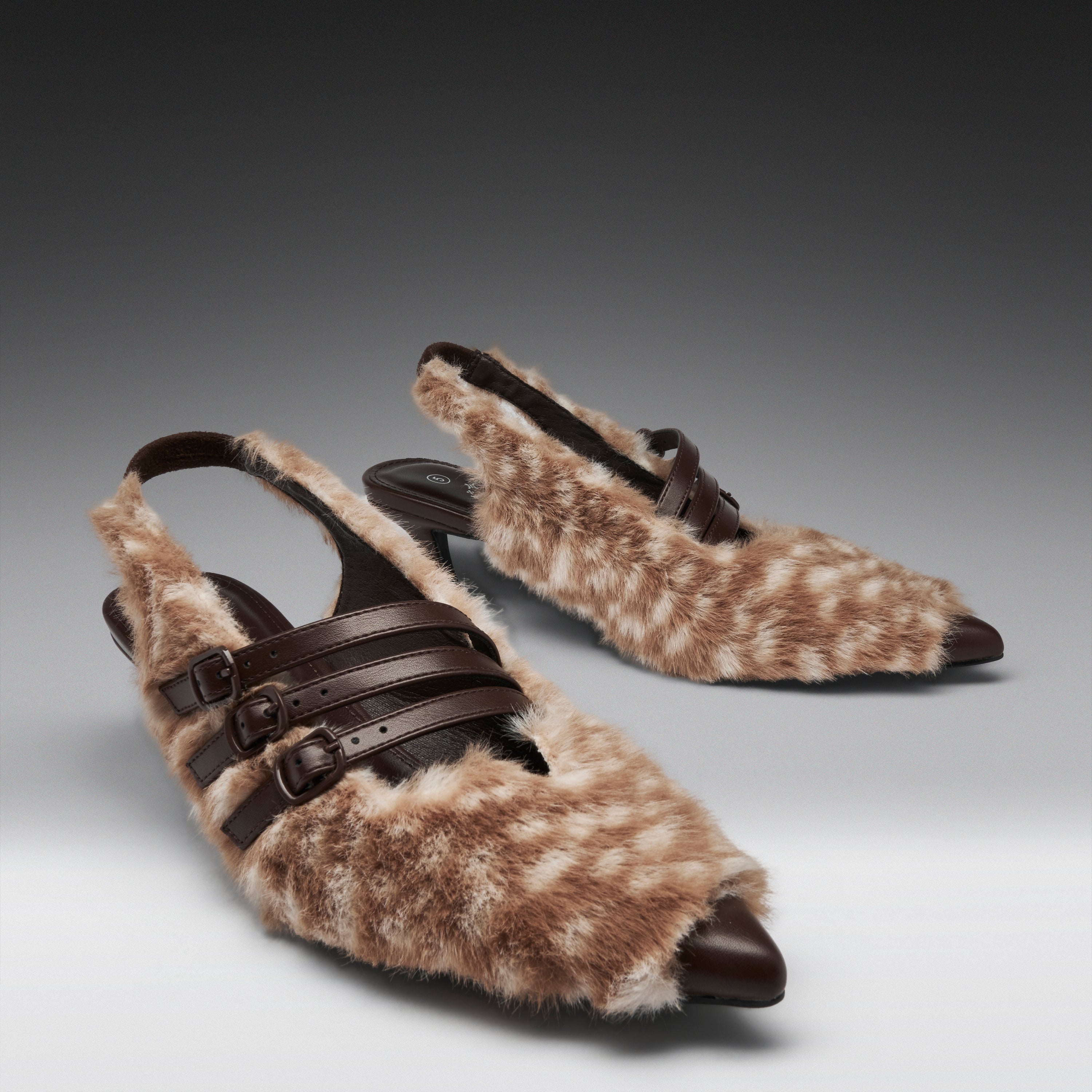 A pair of pointed-toe slingback kitten heels covered in brown and beige spotted faux fur, with dark brown leather straps and buckle details, photographed on a dark-to-light gray gradient studio background.