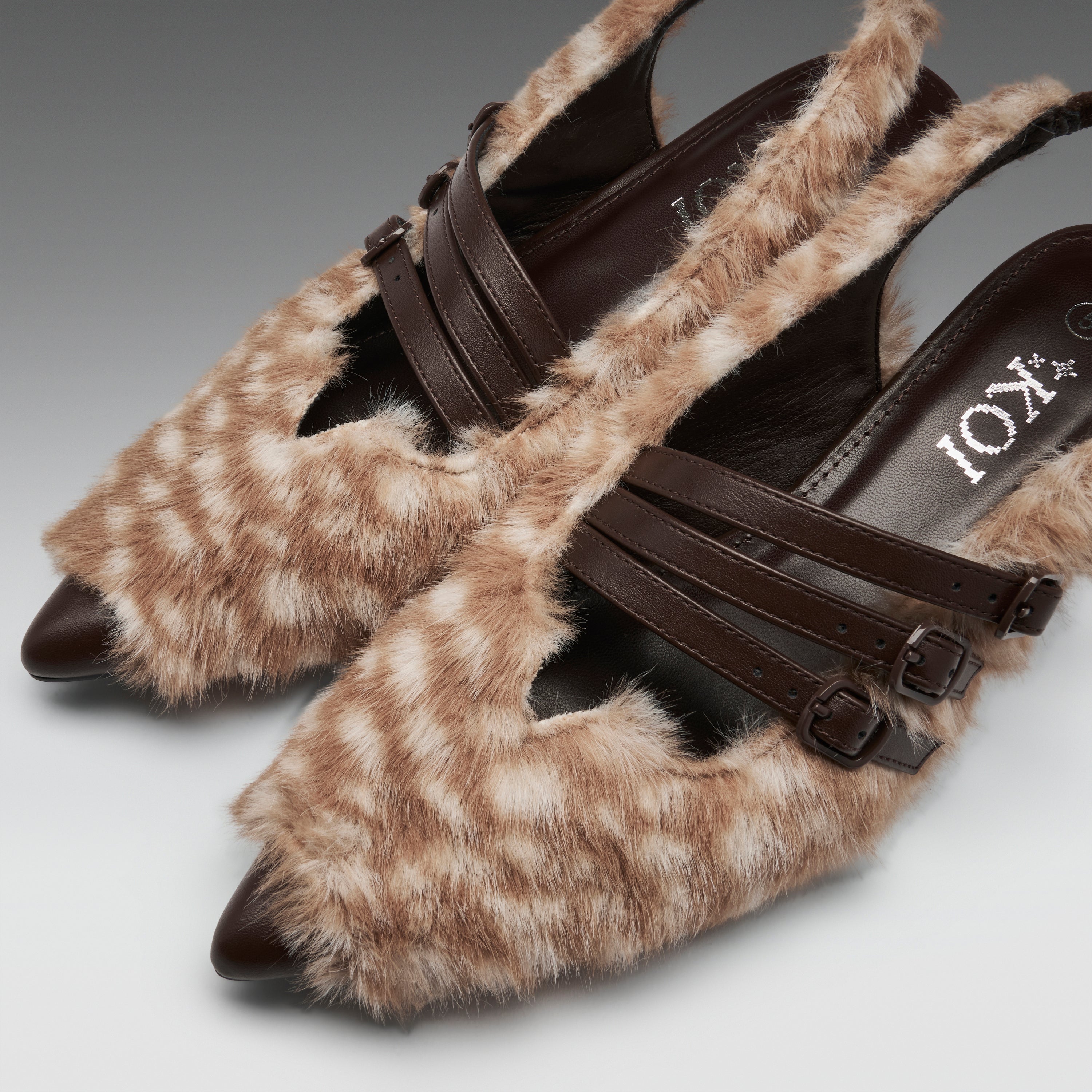 A pair of pointed-toe mule heels covered in a beige and brown spotted faux-fur pattern, with dark brown straps and buckle details across the top. The brand name 'IOX' is visible on the insole.
