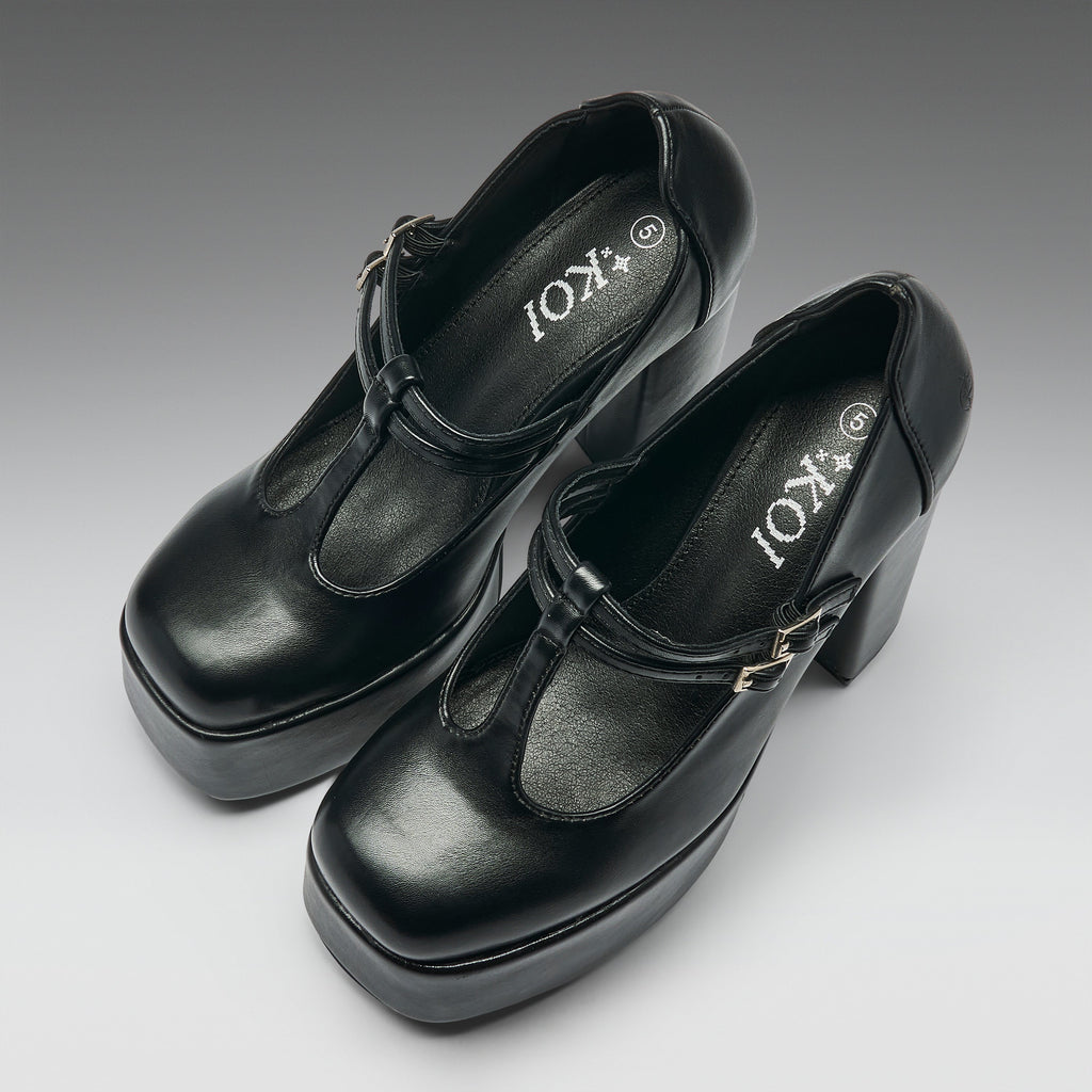 A pair of black leather Mary Jane platform heels with block heels and buckle straps, shown on a neutral gray gradient background. The brand name 'IOKI' is printed on the insoles.