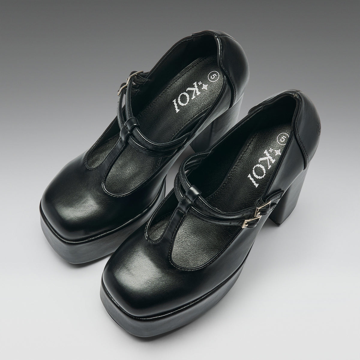 A pair of black leather Mary Jane platform heels with block heels and buckle straps, shown on a neutral gray gradient background. The brand name 'IOKI' is printed on the insoles.