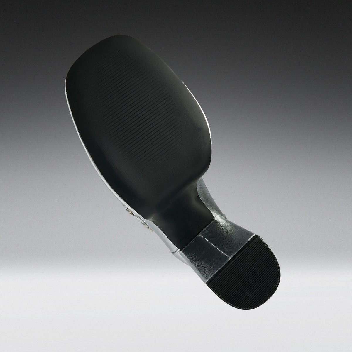 A close-up studio shot of the underside of a black athletic shoe, showing the textured outsole, heel, and a small section of the metallic outsole edge against a gray gradient background.