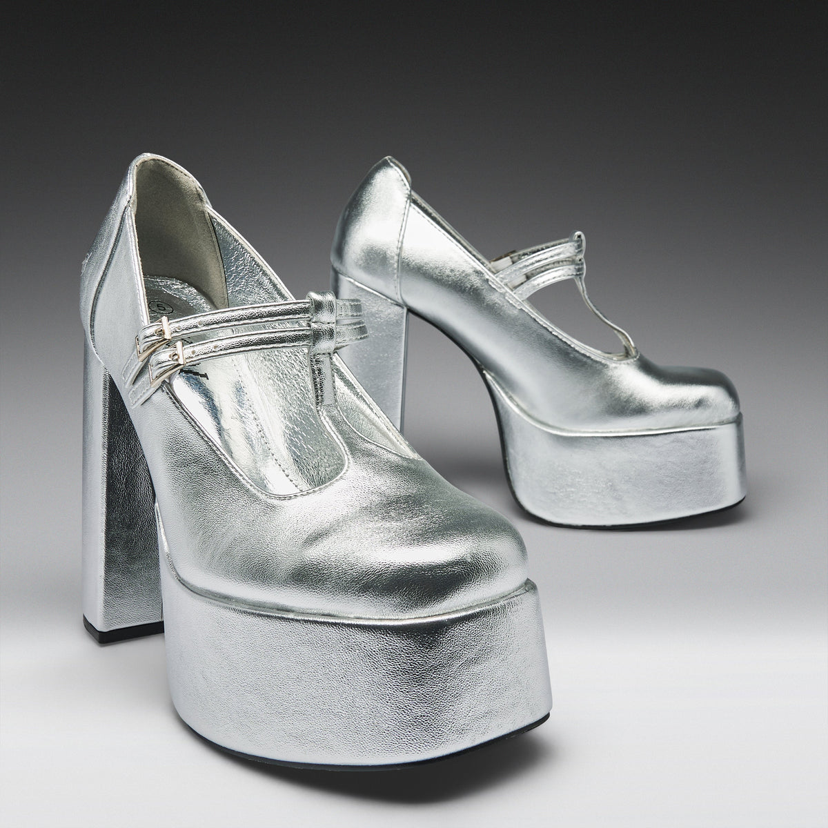 A pair of metallic silver platform Mary Jane high heels with block heels and ankle straps, photographed on a dark-to-light gray gradient studio background.