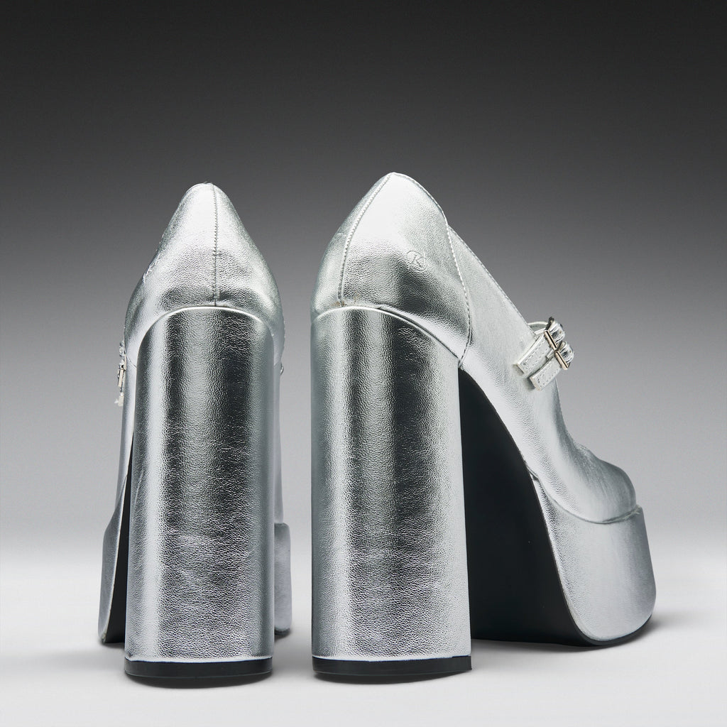 A pair of metallic silver high-heeled pumps with block heels and platform soles, shown from the back on a dark-to-light gray gradient studio background.