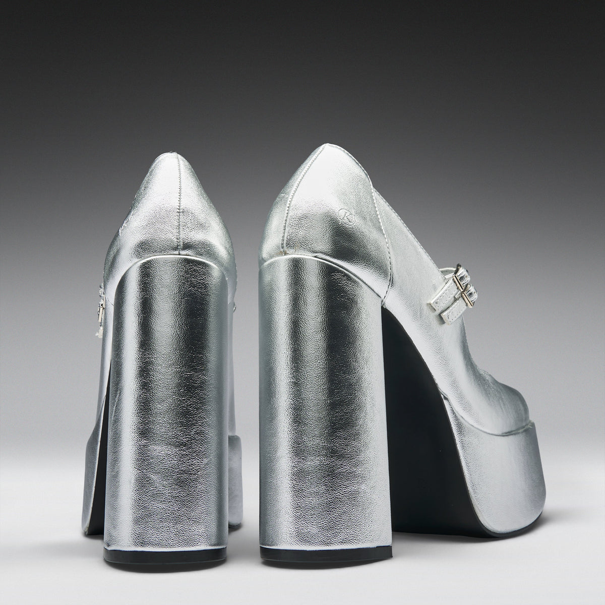 A pair of metallic silver high-heeled pumps with block heels and platform soles, shown from the back on a dark-to-light gray gradient studio background.