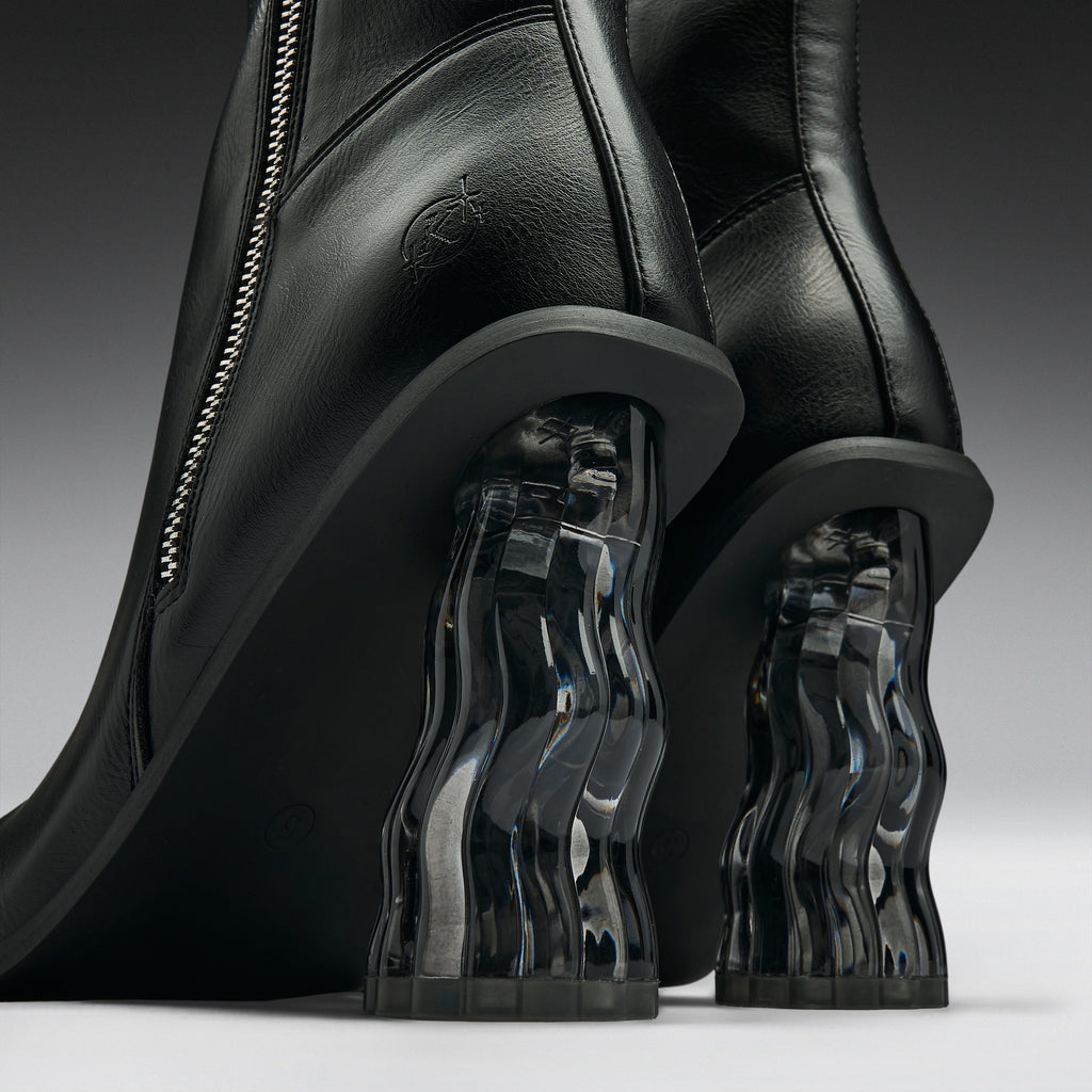 Close-up of a pair of black leather ankle boots with sculptural, wavy transparent acrylic block heels on a dark gray studio background.