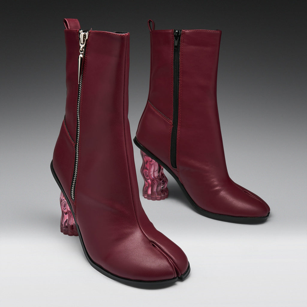 A pair of dark burgundy leather ankle boots with a split toe, side zippers, and chunky translucent pink block heels, shown on a gray gradient studio background.