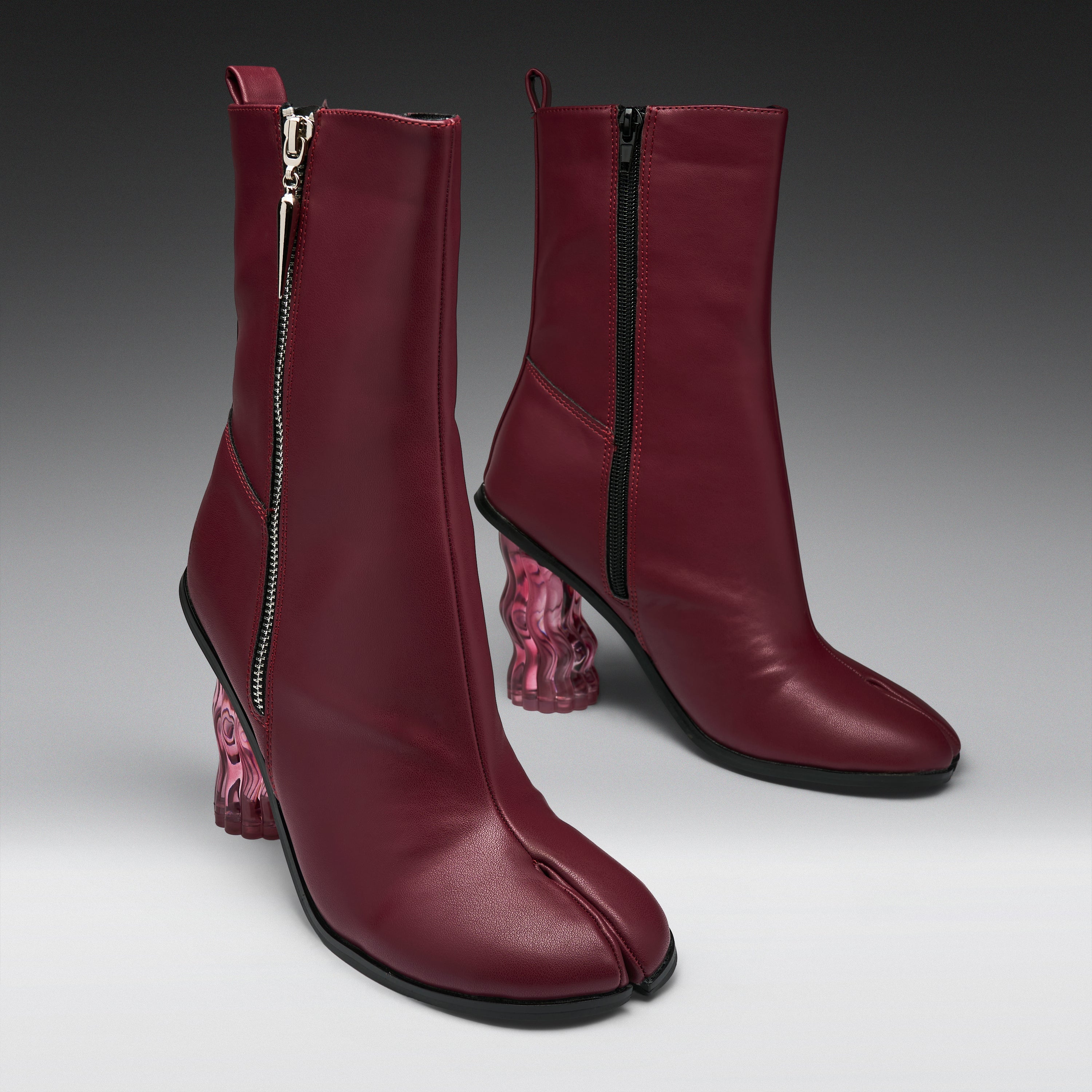 A pair of dark burgundy leather ankle boots with a split toe, side zippers, and chunky translucent pink block heels, shown on a gray gradient studio background.