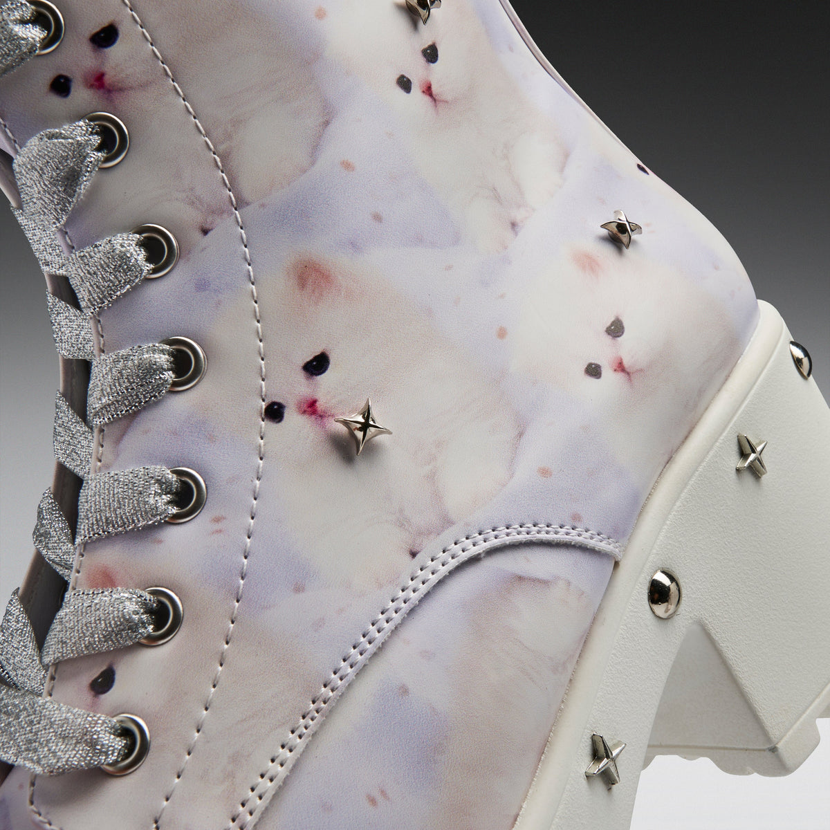 A close-up of a high-top lace-up boot featuring a white and pale lavender watercolor-style print with small pink and black speckles, silver glittery laces, metal eyelets, and star-shaped studs on a white block heel.