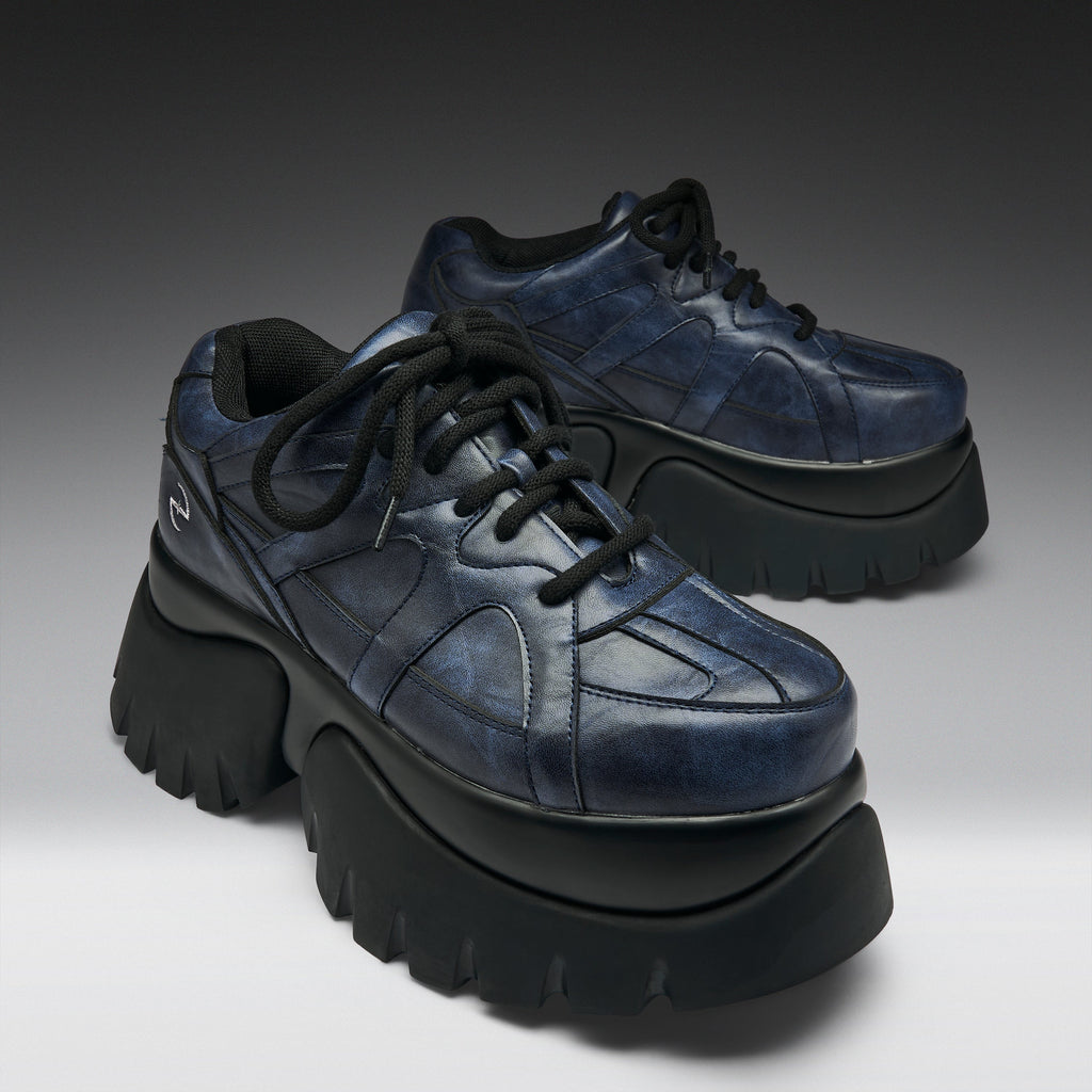 A pair of chunky low-top platform sneakers in distressed navy blue leather with thick black platform soles and black laces, photographed on a dark-to-light gray gradient studio background.