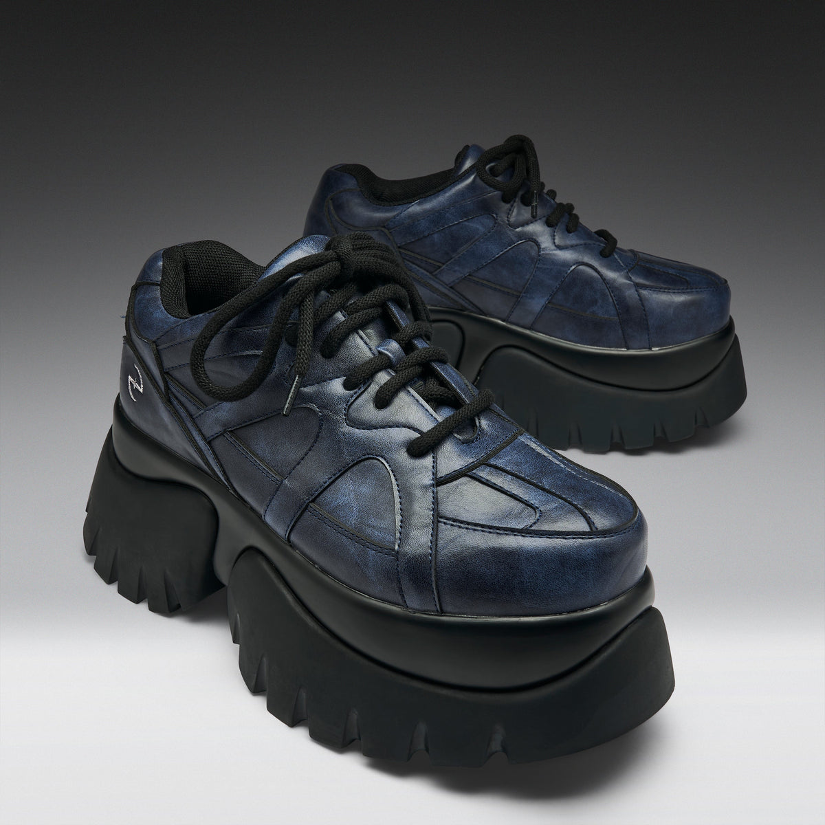 A pair of chunky low-top platform sneakers in distressed navy blue leather with thick black platform soles and black laces, photographed on a dark-to-light gray gradient studio background.