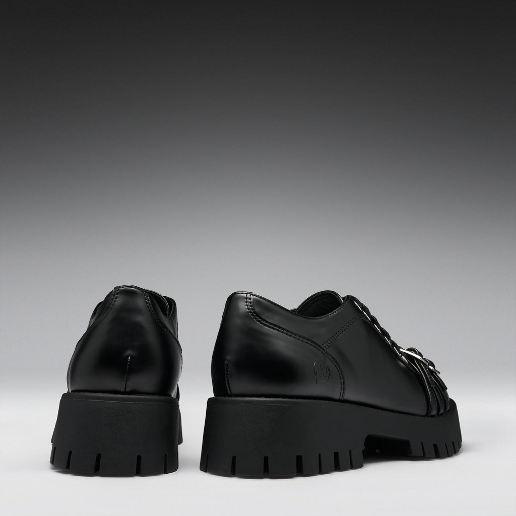 A pair of black leather lace-up shoes with chunky lug soles, shown from the back and side against a dark gray gradient background.