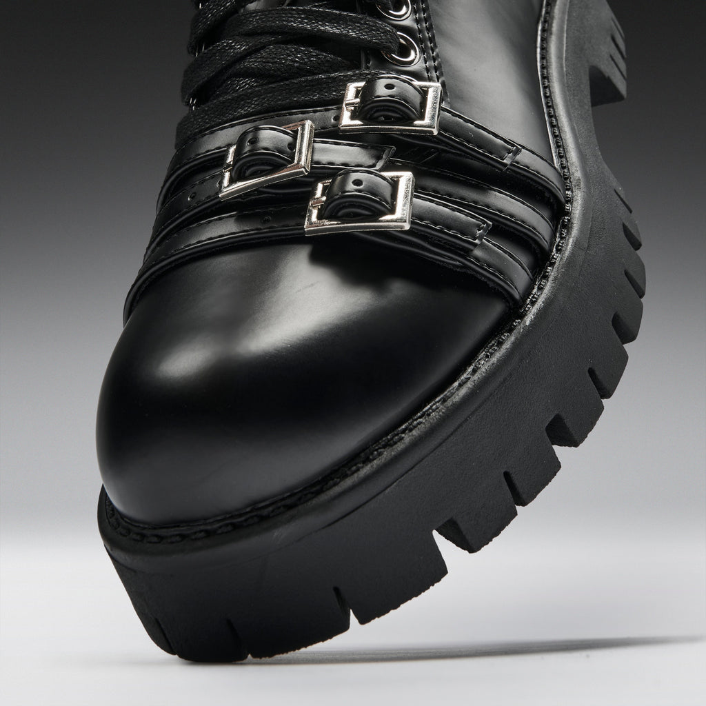 Close-up of a black leather lace-up boot with two silver buckle straps and a chunky lug sole, photographed on a gray studio background.