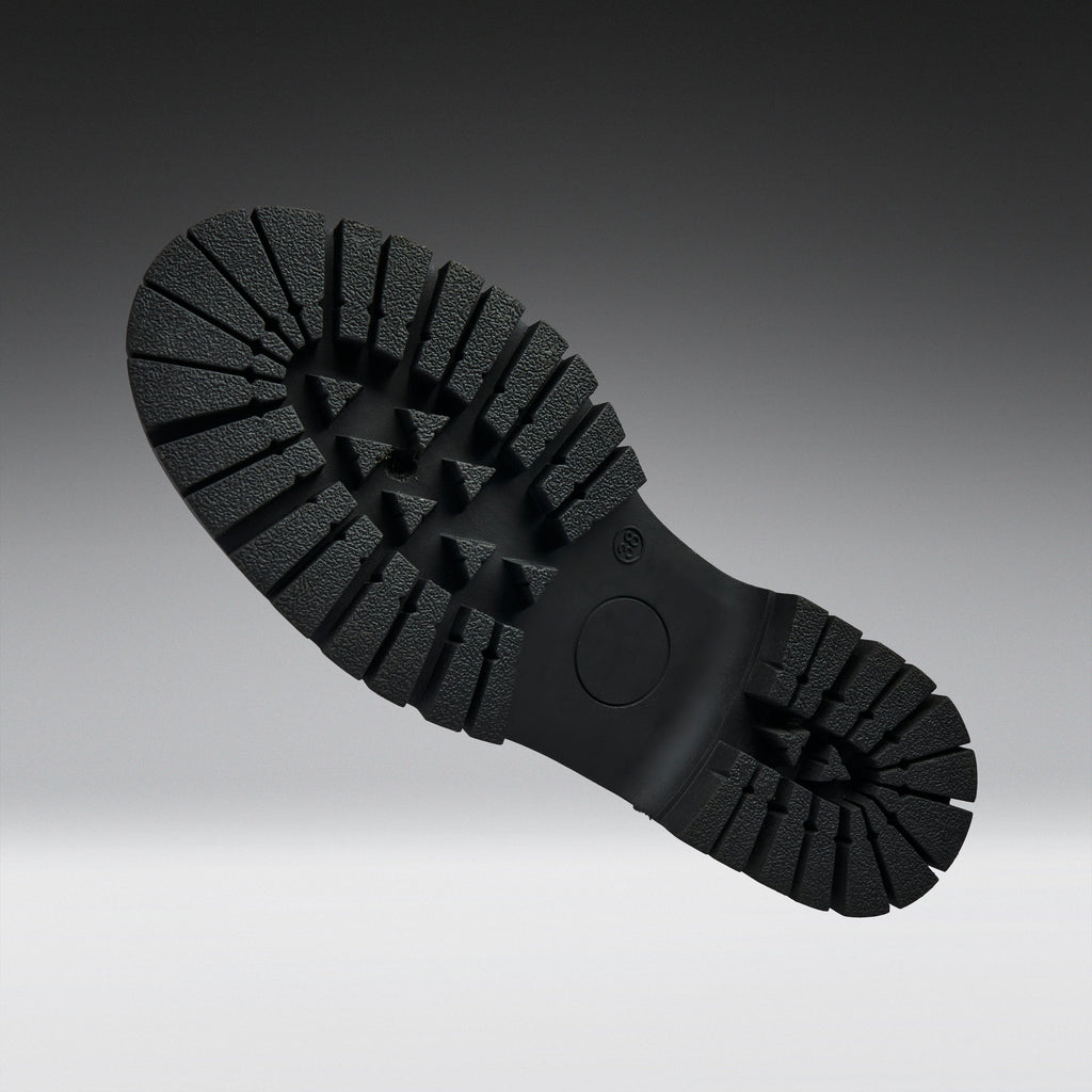 A close-up of a black rubber shoe sole with deep lugs and a circular heel area, photographed against a dark-to-light gray gradient background.