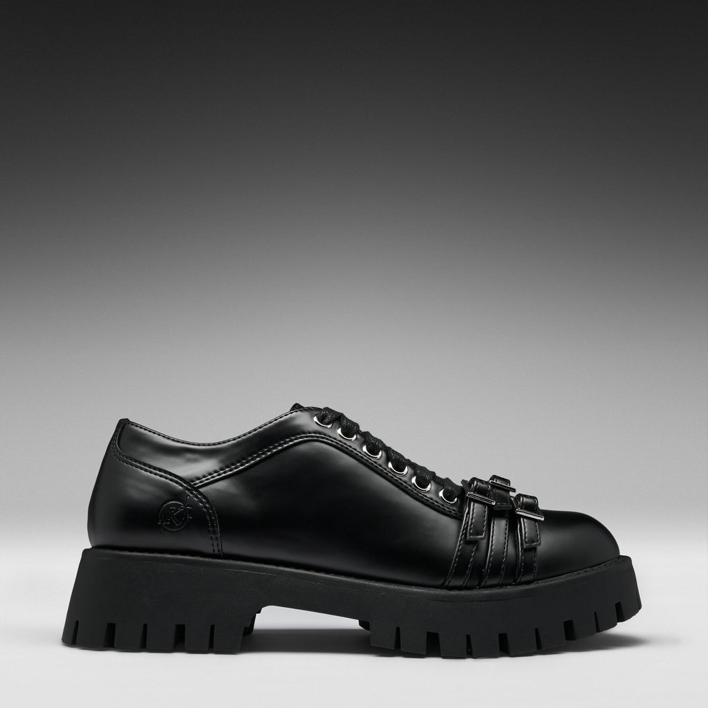 A black leather lace-up shoe with a chunky lug sole and decorative strap details on the toe, shown in a side profile against a dark gray gradient background.