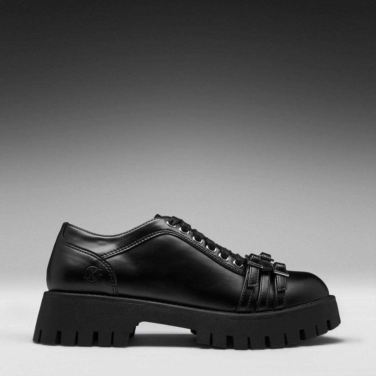 A black leather lace-up shoe with a chunky lug sole and decorative strap details on the toe, shown in a side profile against a dark gray gradient background.