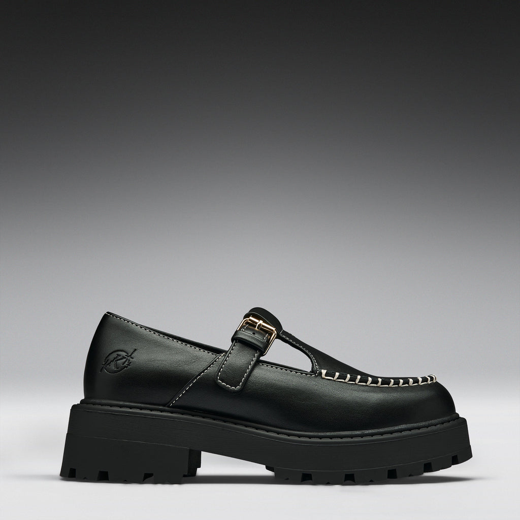 A single black leather Mary Jane loafer with a chunky lug sole, a gold buckle strap, and decorative gold stitching on the toe, shown against a dark gray gradient background.