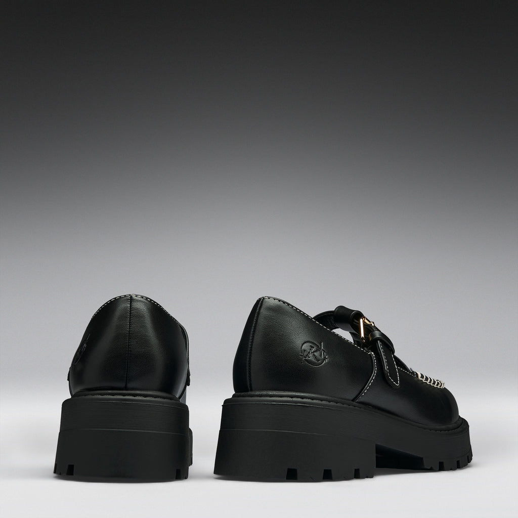 A pair of black leather platform mule shoes shown from the back, featuring chunky lug soles, double straps, and a small gold-tone buckle detail on a dark gray gradient background.