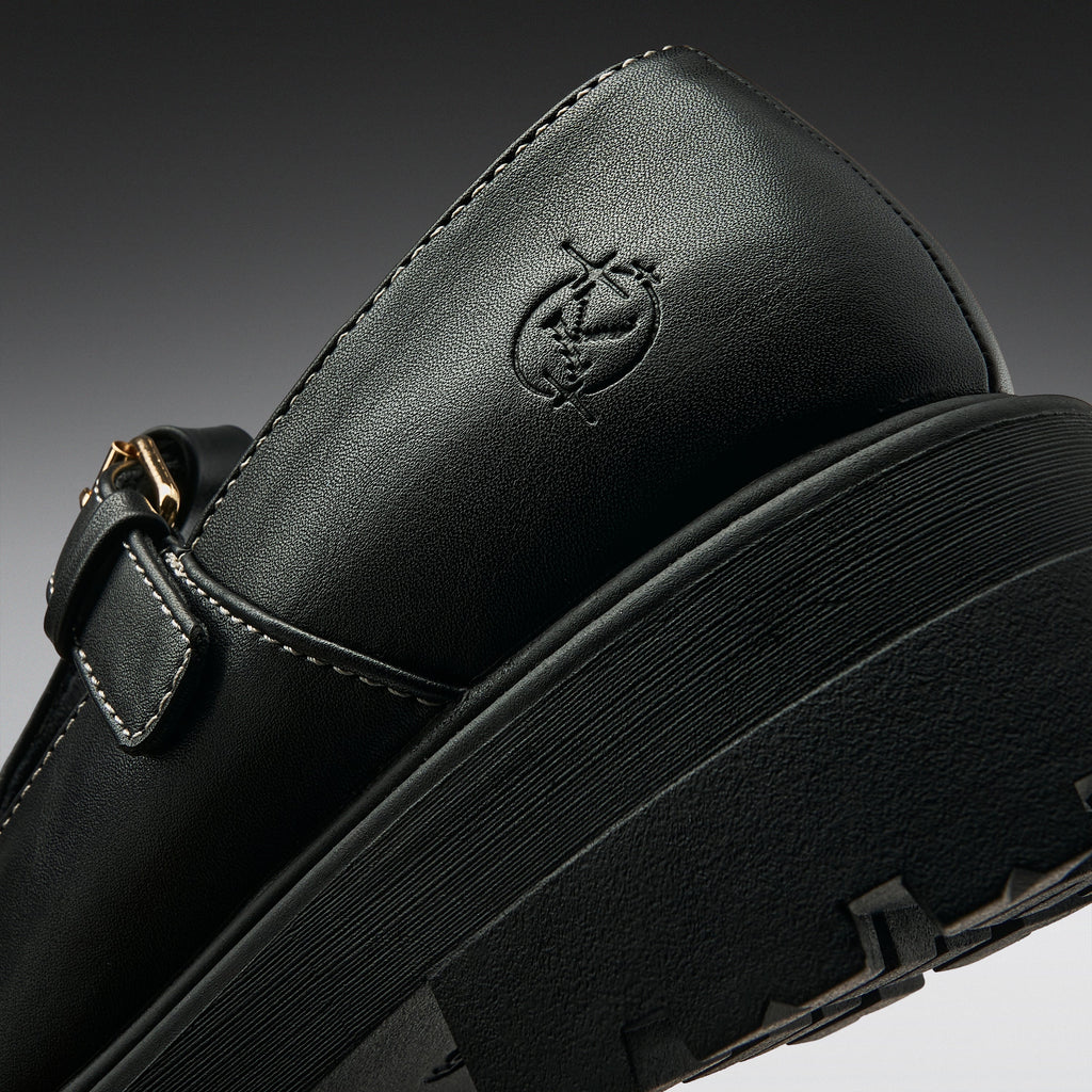 Close-up of a black leather Mary Jane shoe with a thick black platform sole, a side buckle strap, and an embossed logo on the heel, shown against a dark gray gradient background.