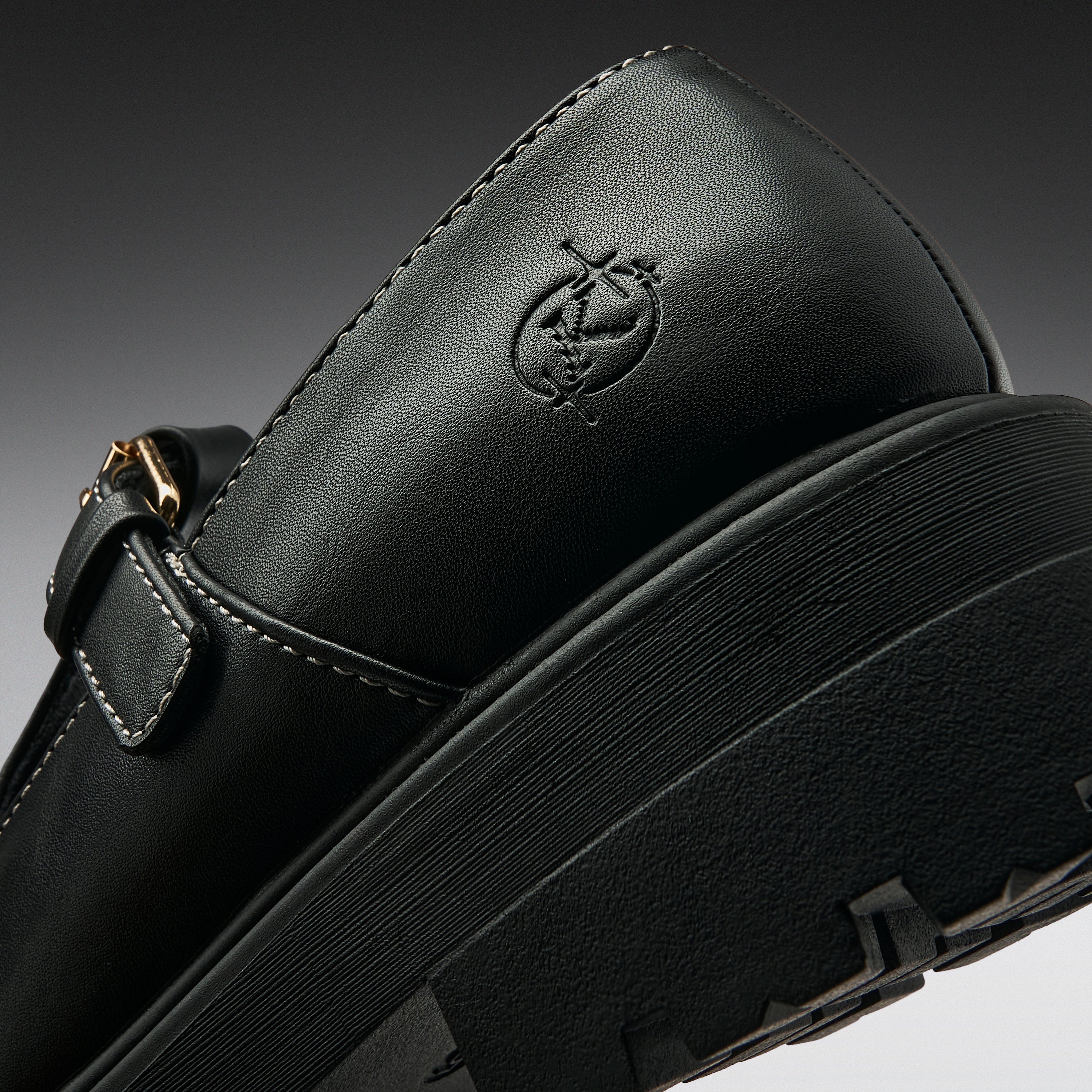 Close-up of a black leather Mary Jane shoe with a thick black platform sole, a side buckle strap, and an embossed logo on the heel, shown against a dark gray gradient background.