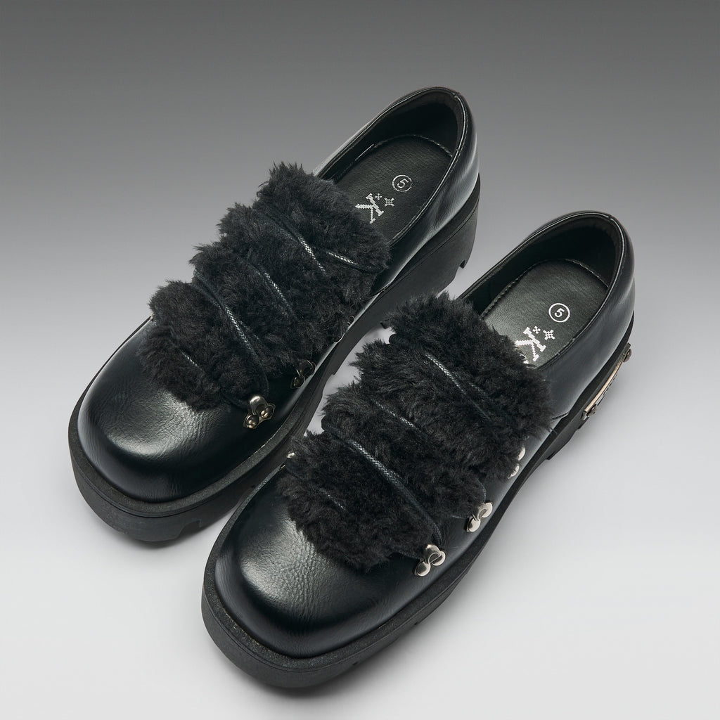 A pair of black leather platform loafers with wide, fluffy black faux-fur straps and small silver metal accents, photographed on a neutral gray gradient background.