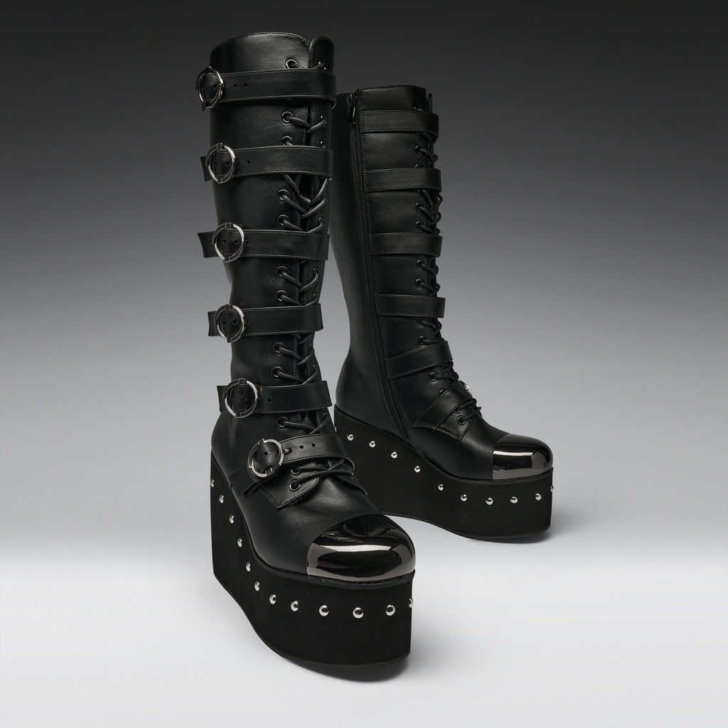 A pair of black platform knee-high boots with multiple buckle straps, lace-up front, and silver studded details, photographed on a dark-to-light gray gradient studio background.