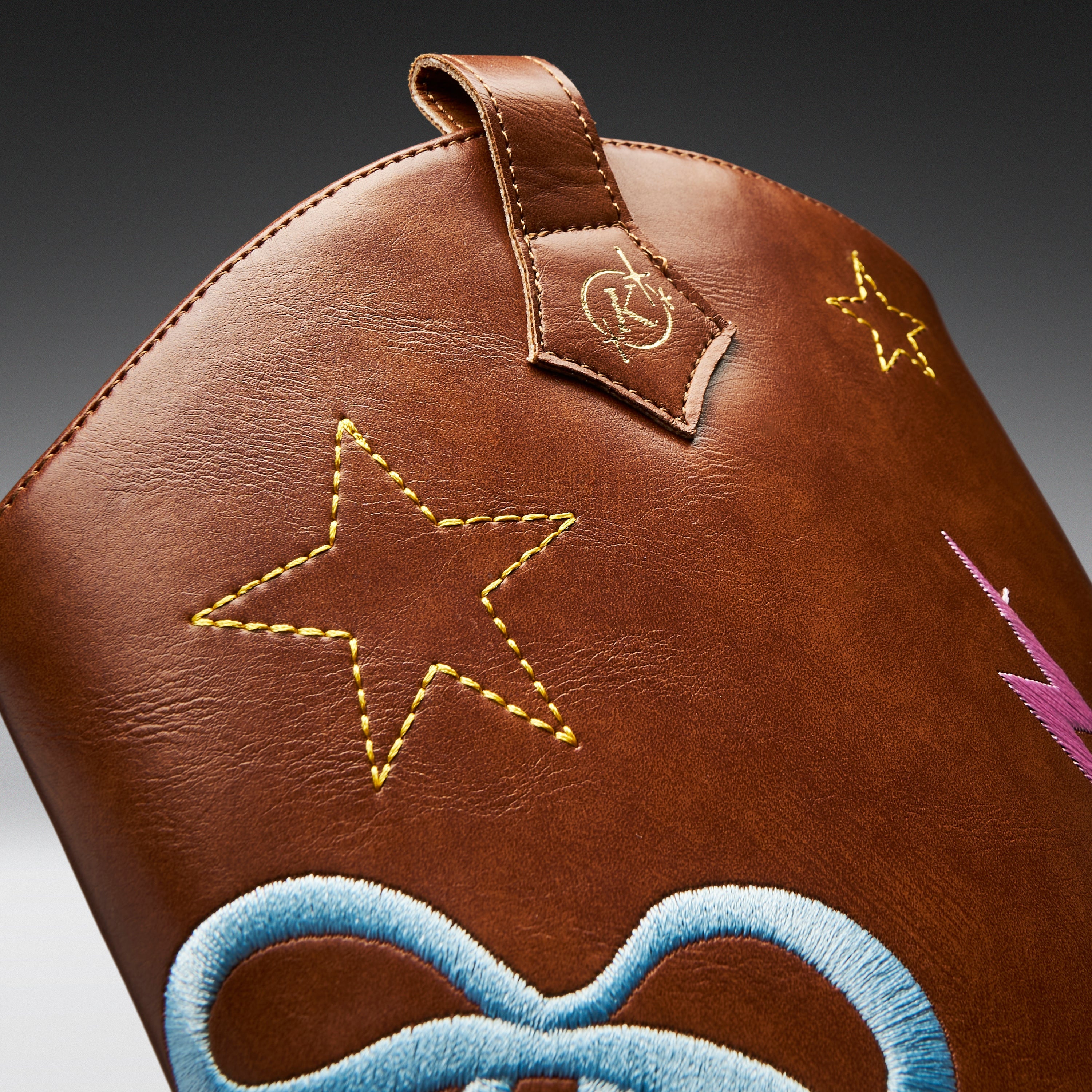 Close-up of a brown leather bag featuring embroidered star motifs in yellow, a blue bow design at the bottom, and a small pink lightning bolt detail on a dark gradient background.