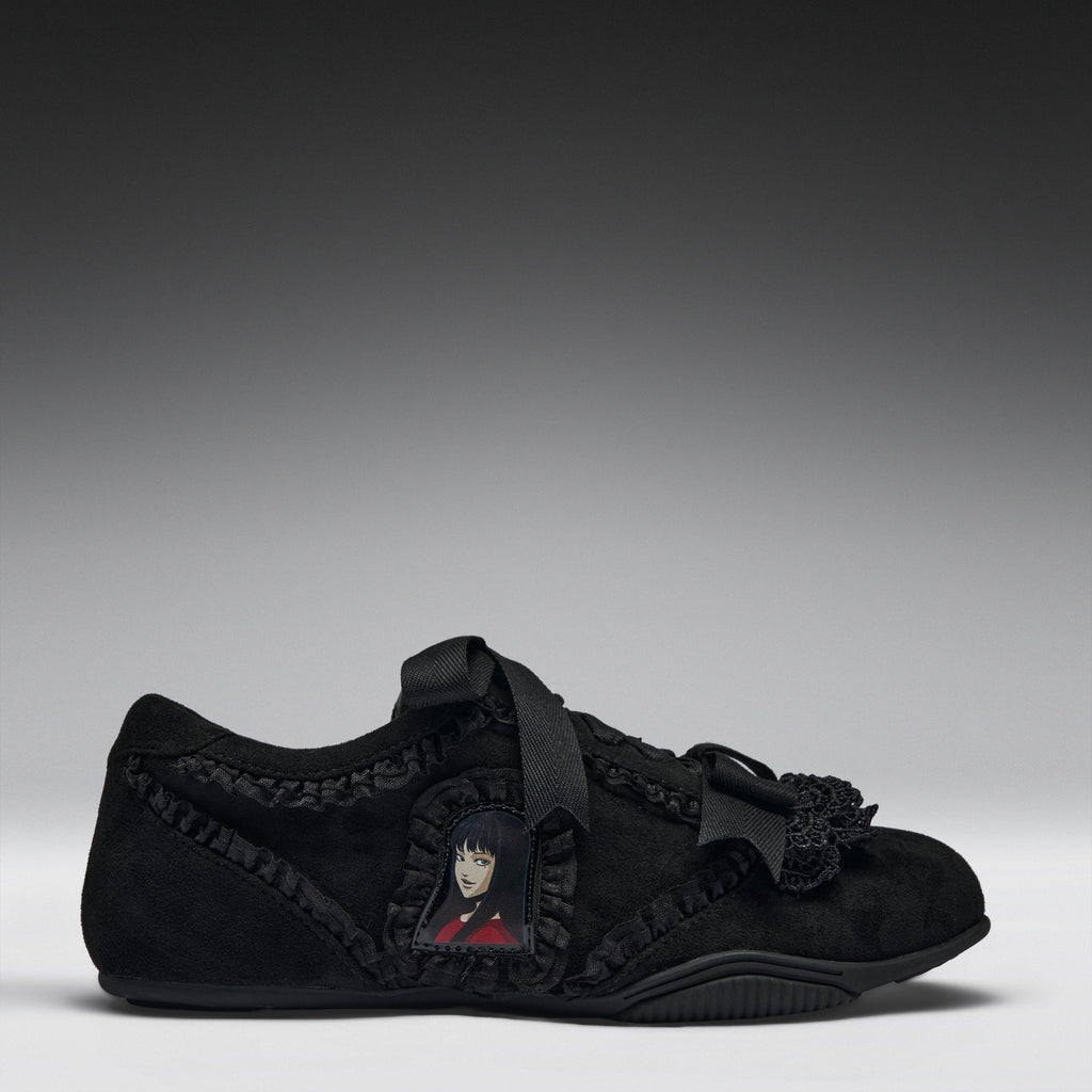 A black suede slip-on shoe featuring a decorative black ribbon strap, ruffled trim, a black tassel accent, and a small anime character patch on the side, set against a dark gray gradient background.
