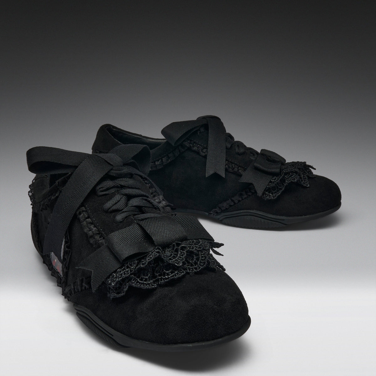 A pair of black lace-up shoes with wide black ribbon bows, black lace trim, and textured black uppers, photographed on a dark-to-light gray studio background.