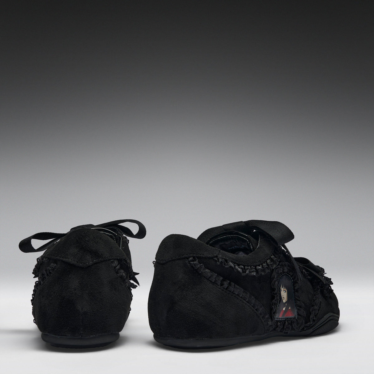 A pair of black suede baby moccasins with fringed trim and lace-up front details, shown from the back and side against a dark gray gradient background.