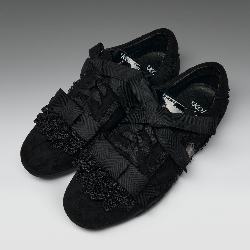 A pair of black lace-up flats with wide satin ribbon straps across the toes and decorative black lace trim on the vamp, photographed on a neutral gray studio background.