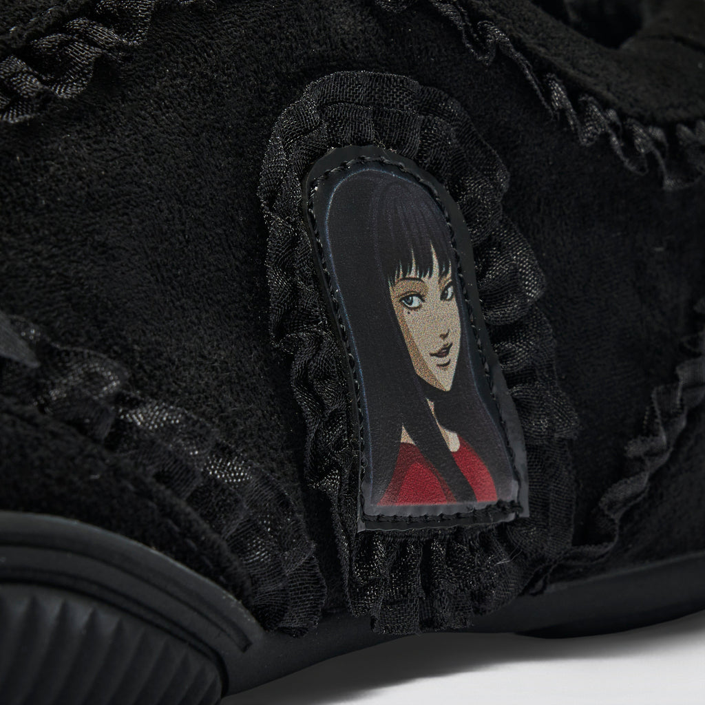 Close-up of a black suede sneaker featuring a ruffled lace detail and a central patch with an illustrated anime-style character with black hair and a red top.