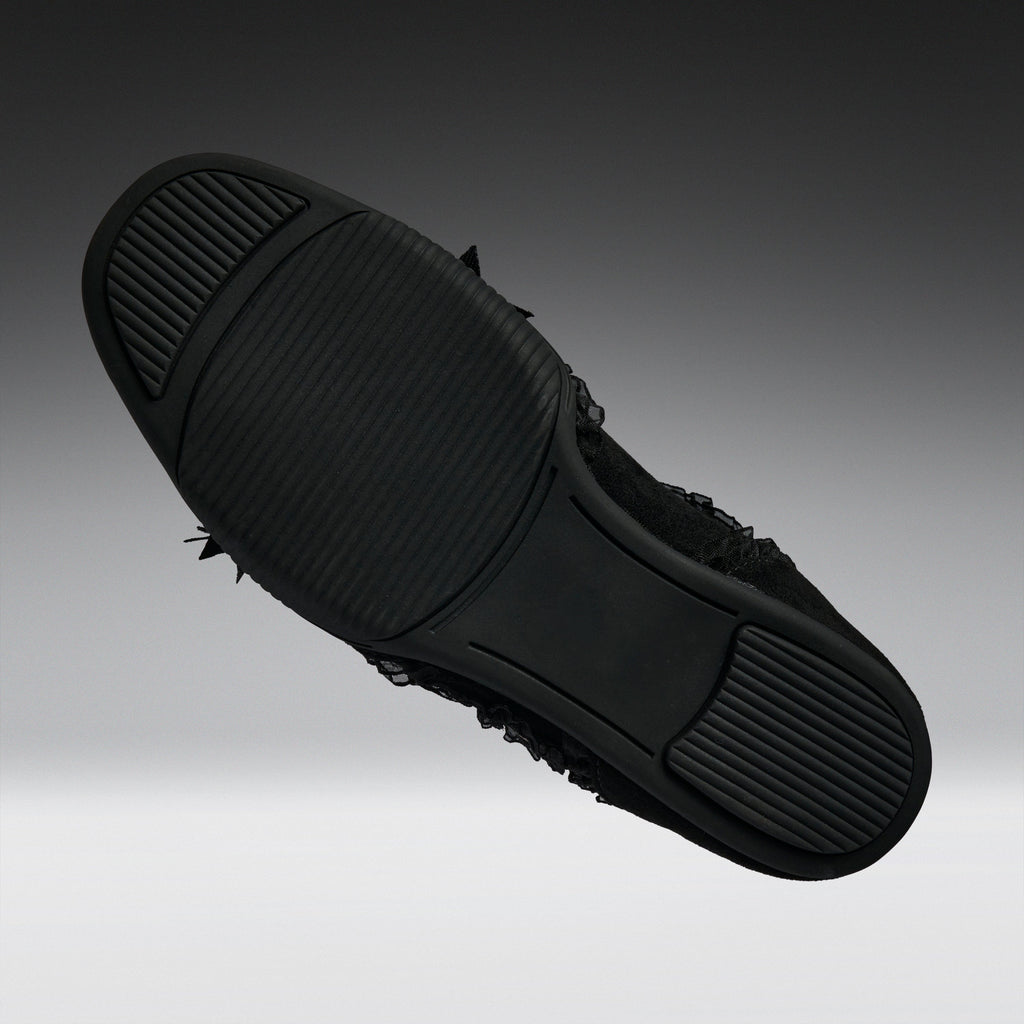 A black slip-on shoe shown from a top-down angle, highlighting its textured rubber sole with small black frayed details along the side, set against a dark gray gradient background.