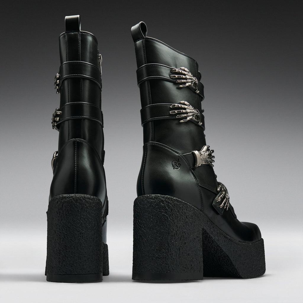 A pair of black leather ankle boots with chunky platform soles and high block heels, decorated with silver skeleton hand and chain buckle embellishments, shown from the back and side against a dark gray gradient background.